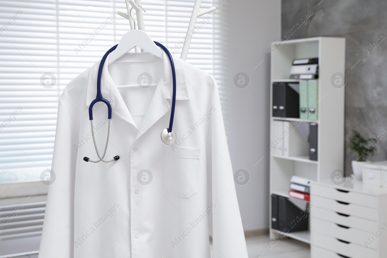 Photo of White doctor's gown and stethoscope hanging on rack in clinic, closeup. Space for text