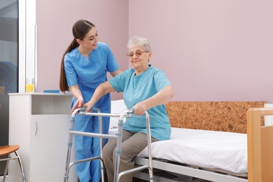 Nurse assisting senior woman with walker to get up from bed in hospital ward