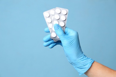 Scientist in protective gloves holding pills on light blue background, closeup