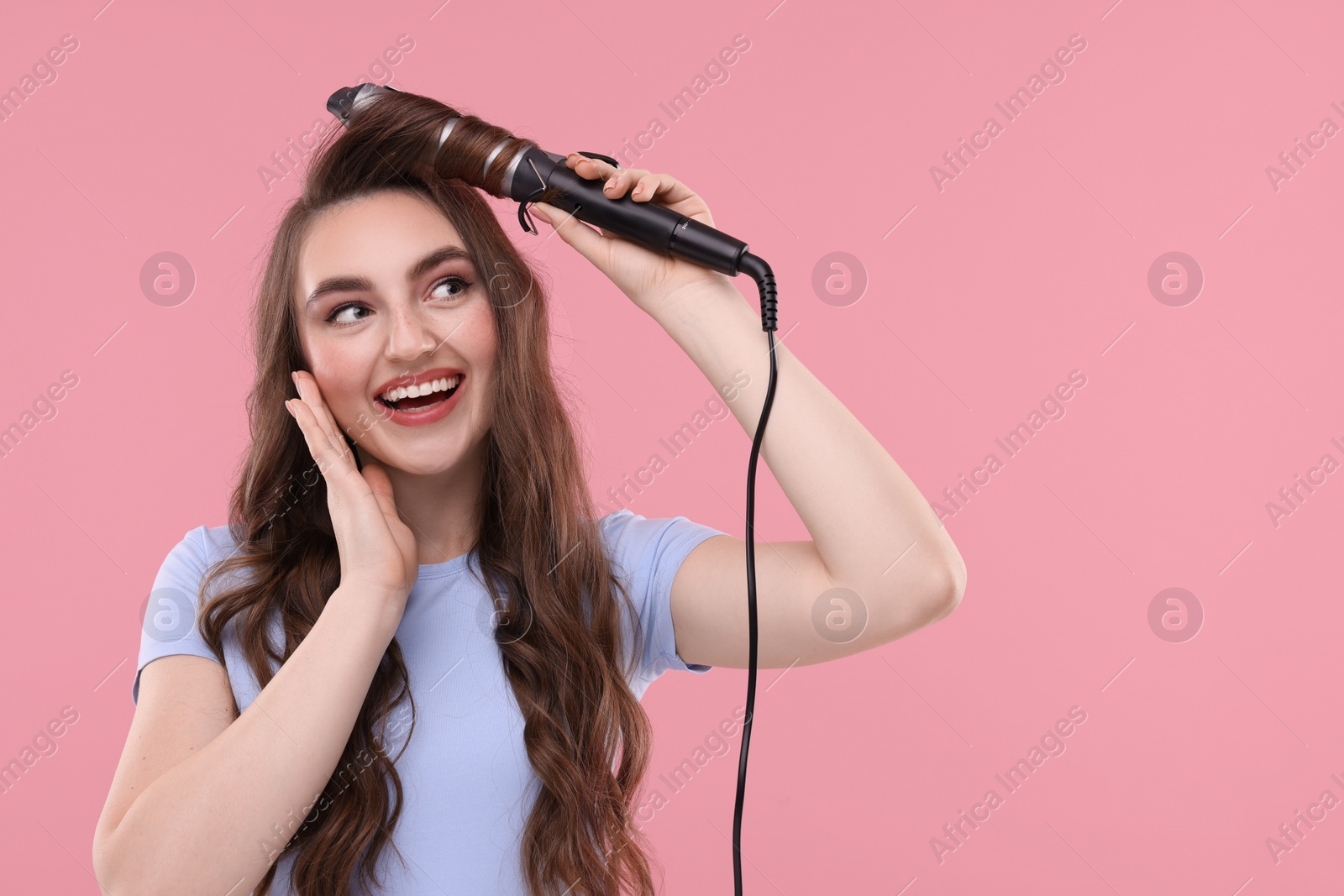 Photo of Beautiful young woman using curling hair iron on pink background, space for text