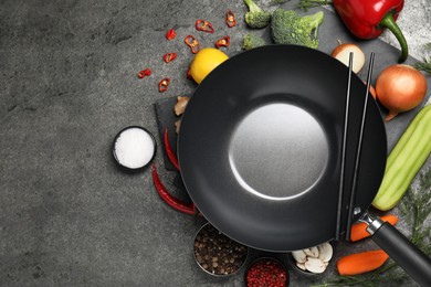 Empty iron wok and chopsticks surrounded by ingredients on grey table, flat lay. Space for text