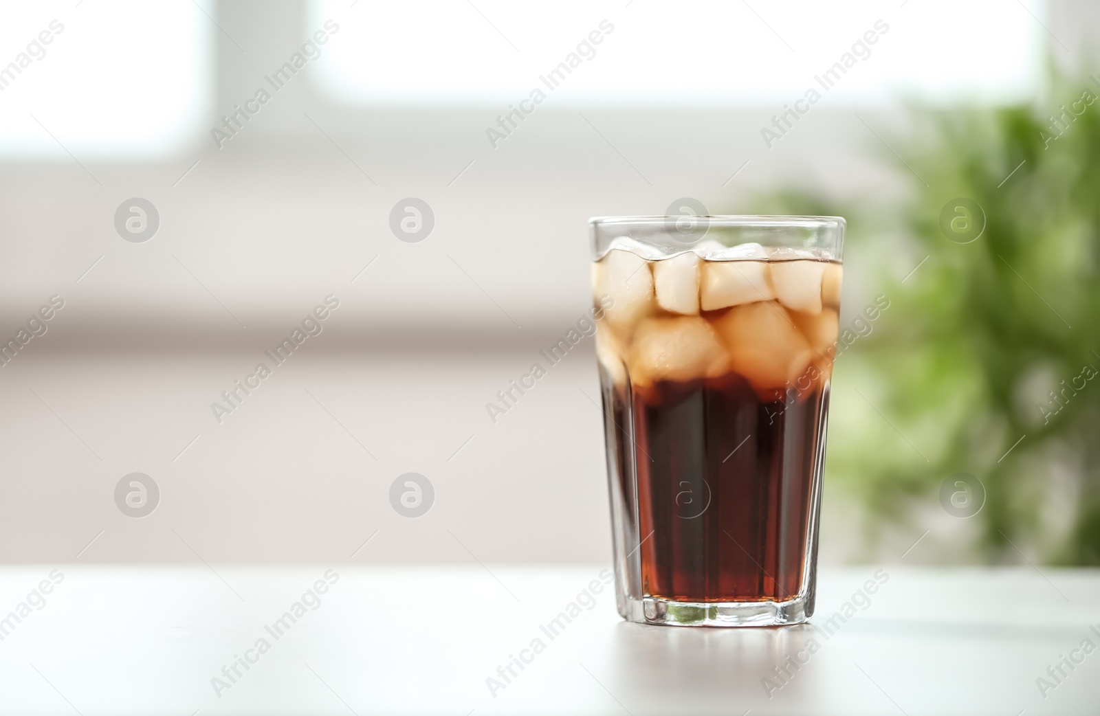 Photo of Glass of cola with ice on table against blurred background, space for text