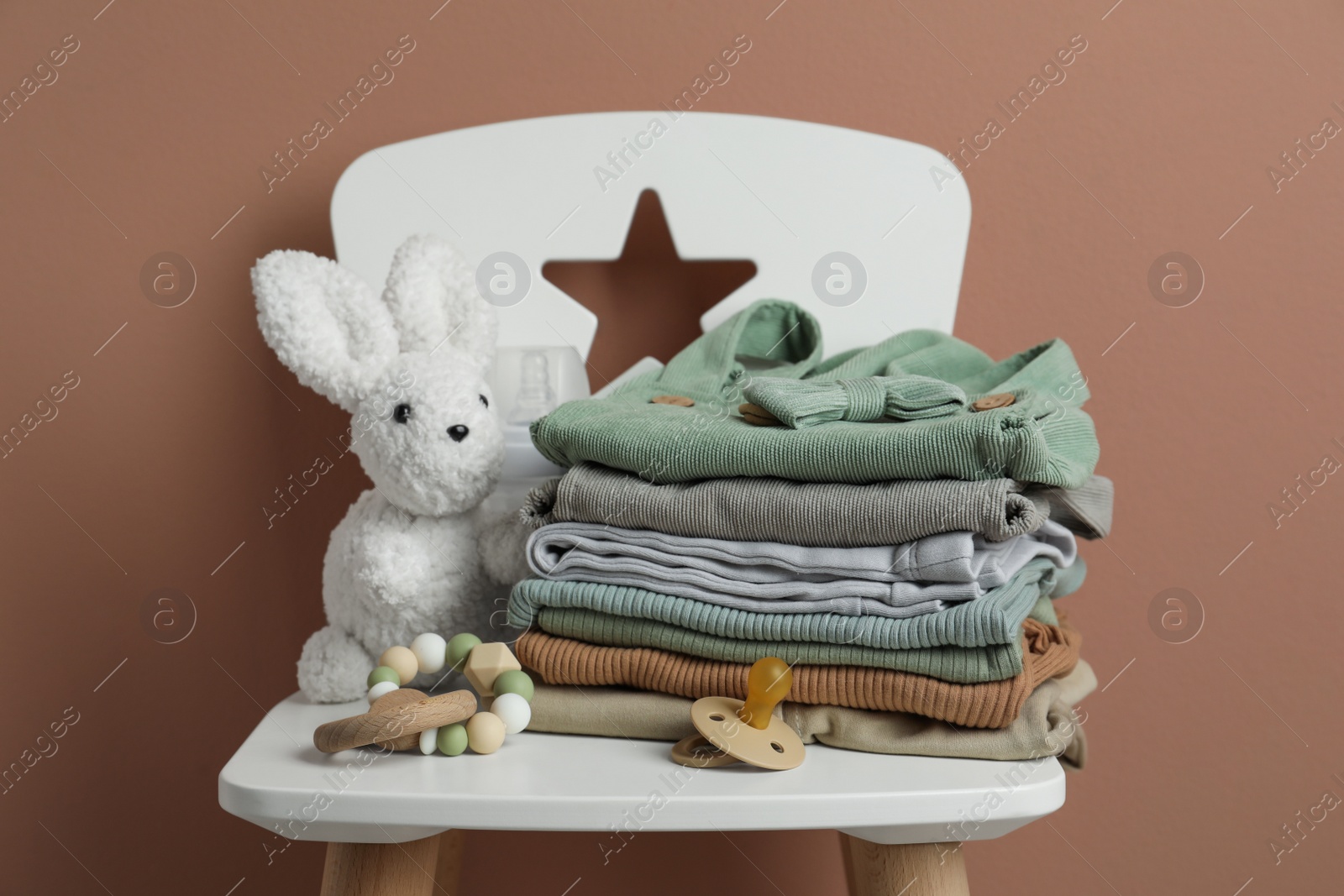 Photo of Stack of baby clothes, pacifier and toys on chair near light brown wall