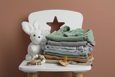 Photo of Stack of baby clothes, pacifier and toys on chair near light brown wall