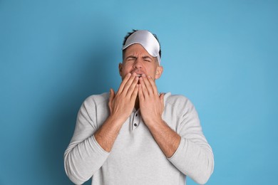 Tired man with sleeping mask yawning on light blue background