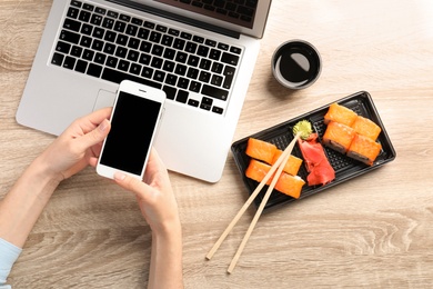 Woman with smartphone, laptop and tasty sushi rolls at wooden table, space for text. Food delivery