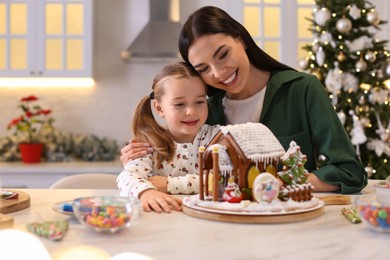 Photo of Mother and daughter with gingerbread house at table indoors