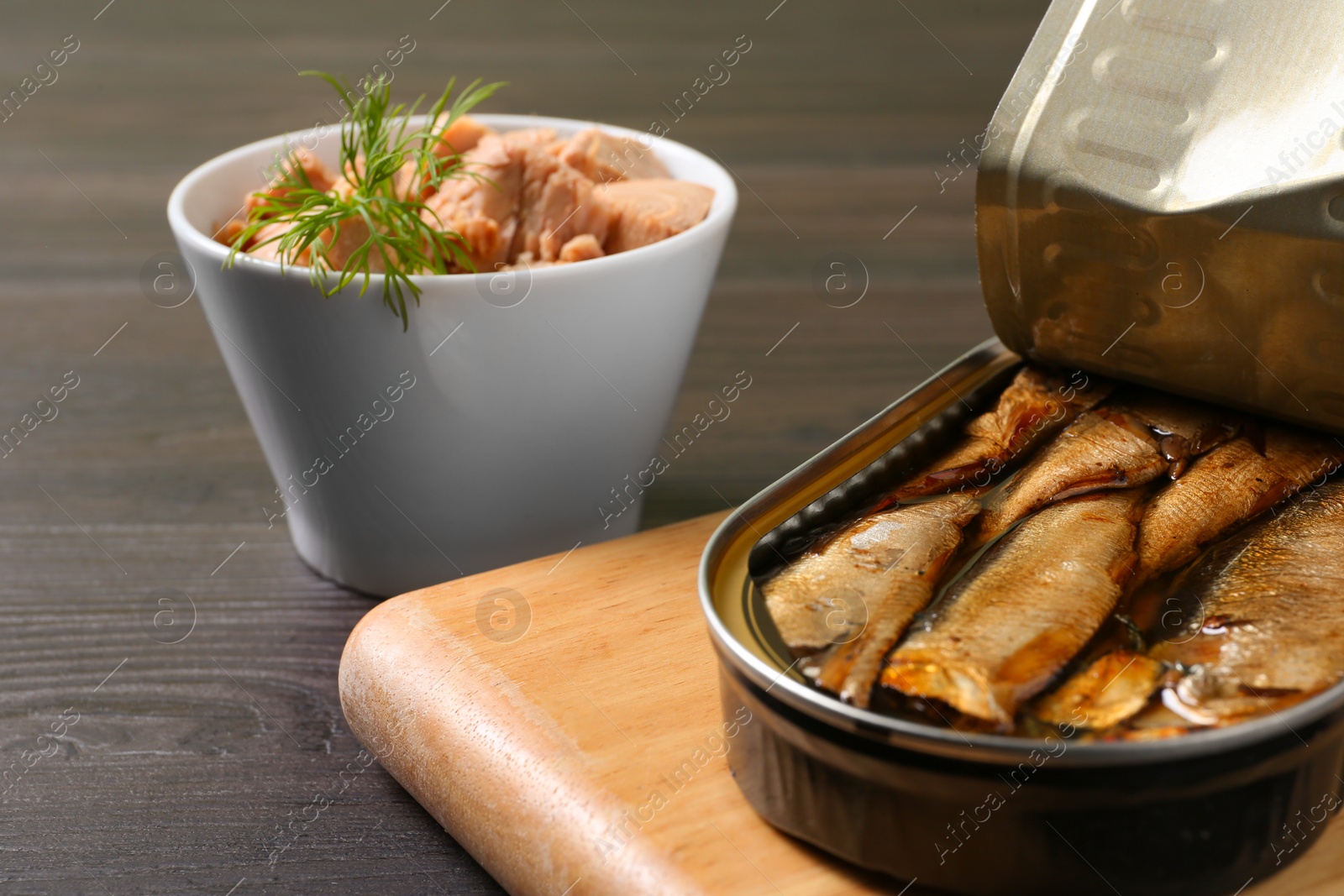 Photo of Board with tin can of sprats and tuna in bowl on wooden table, closeup. Space for text