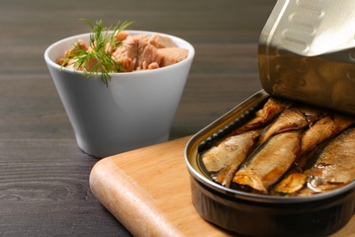 Photo of Board with tin can of sprats and tuna in bowl on wooden table, closeup. Space for text