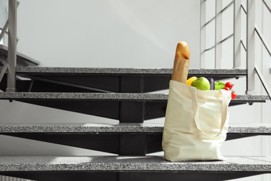 Tote bag with vegetables and bread on stairs indoors. Space for text