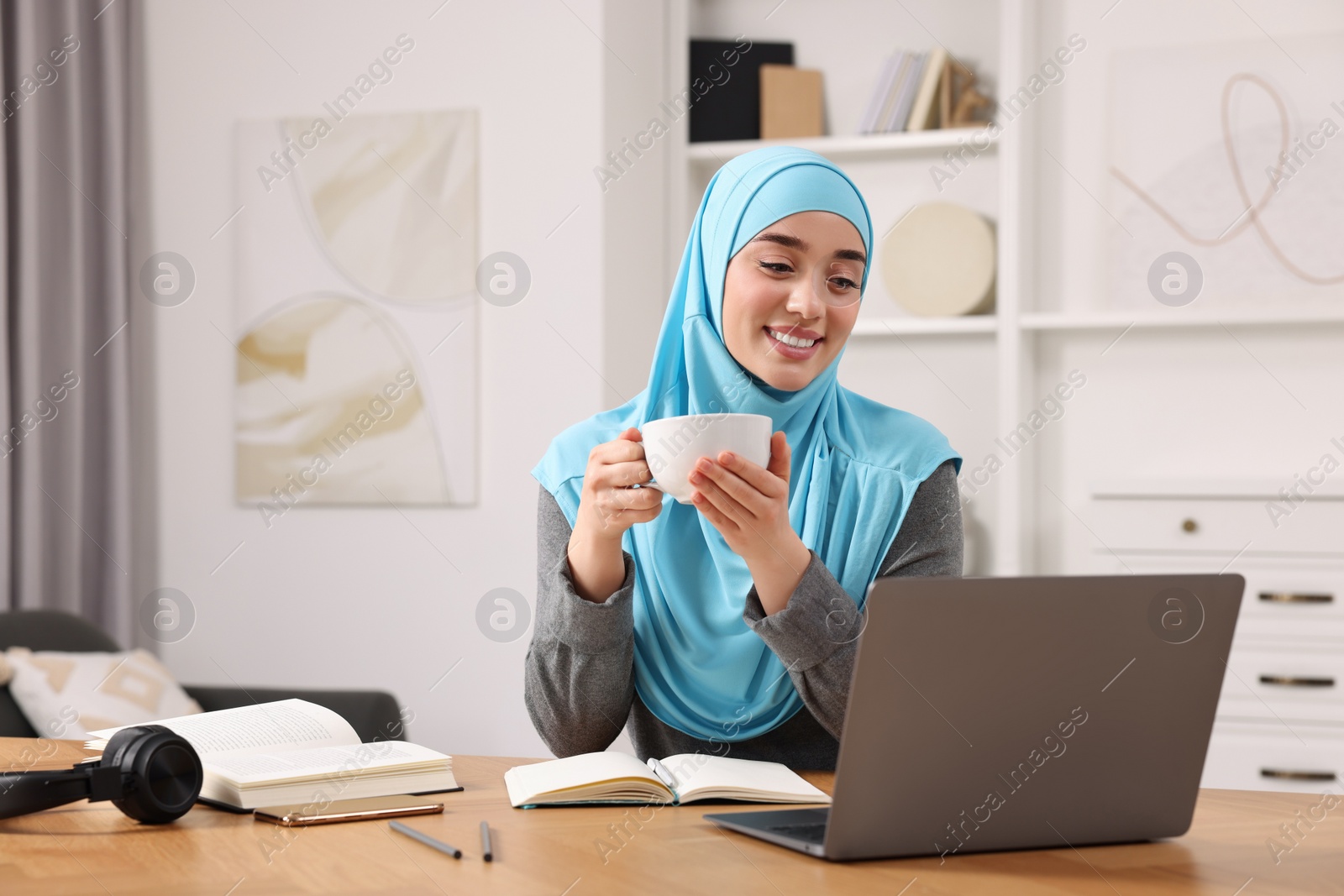 Photo of Muslim woman in hijab with cup of coffee using laptop at wooden table in room