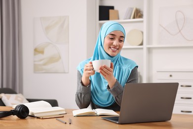 Muslim woman in hijab with cup of coffee using laptop at wooden table in room