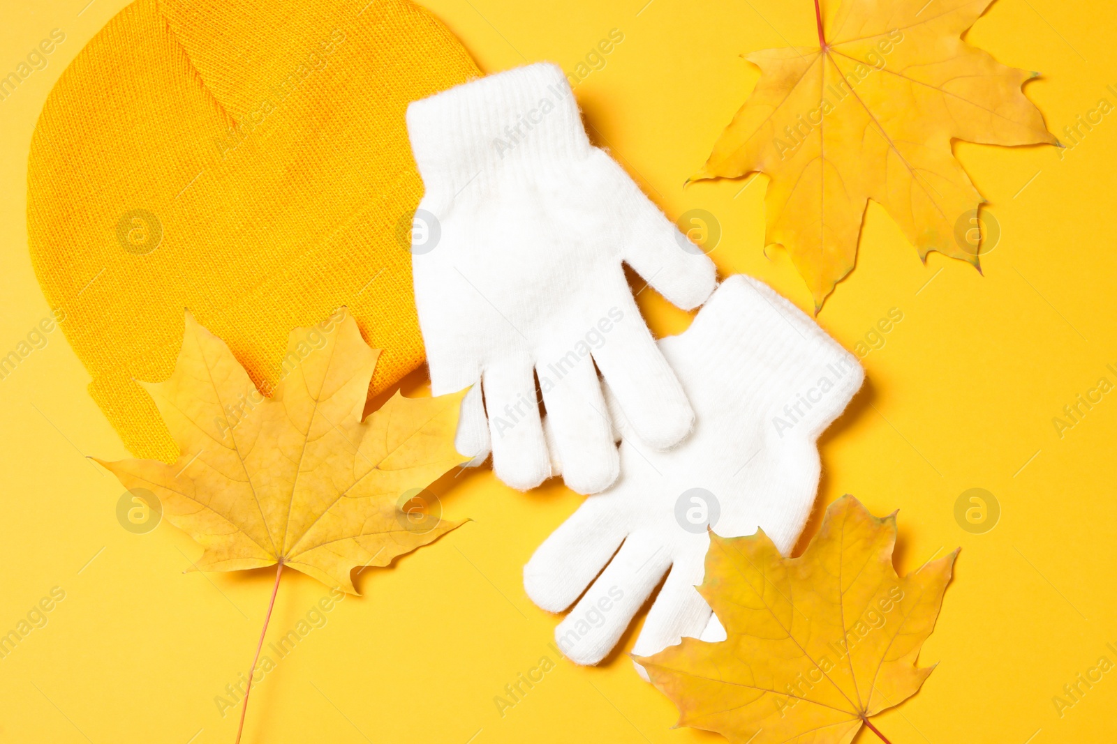 Photo of Stylish white woolen gloves, hat and dry leaves on yellow background, flat lay