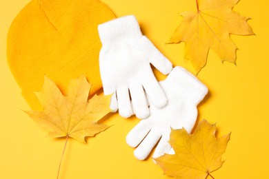 Stylish white woolen gloves, hat and dry leaves on yellow background, flat lay