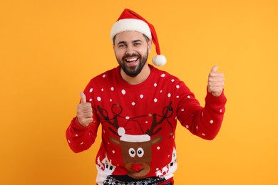 Photo of Happy young man in Christmas sweater and Santa hat showing thumbs up on orange background