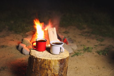 Cups of hot drink on old log near bonfire outdoors. Camping season