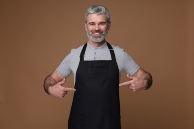 Photo of Happy man pointing at kitchen apron on brown background. Mockup for design