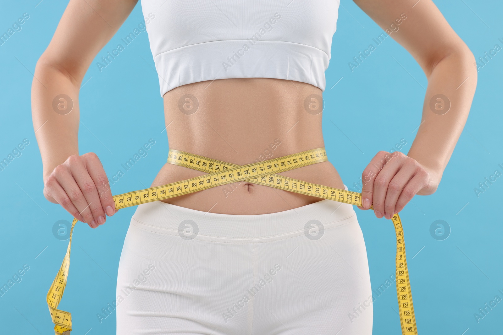 Photo of Slim woman measuring waist with tape on light blue background, closeup. Weight loss