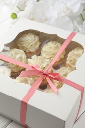 Photo of Tasty cupcakes with vanilla cream in box and orchid flowers on white wooden table, closeup