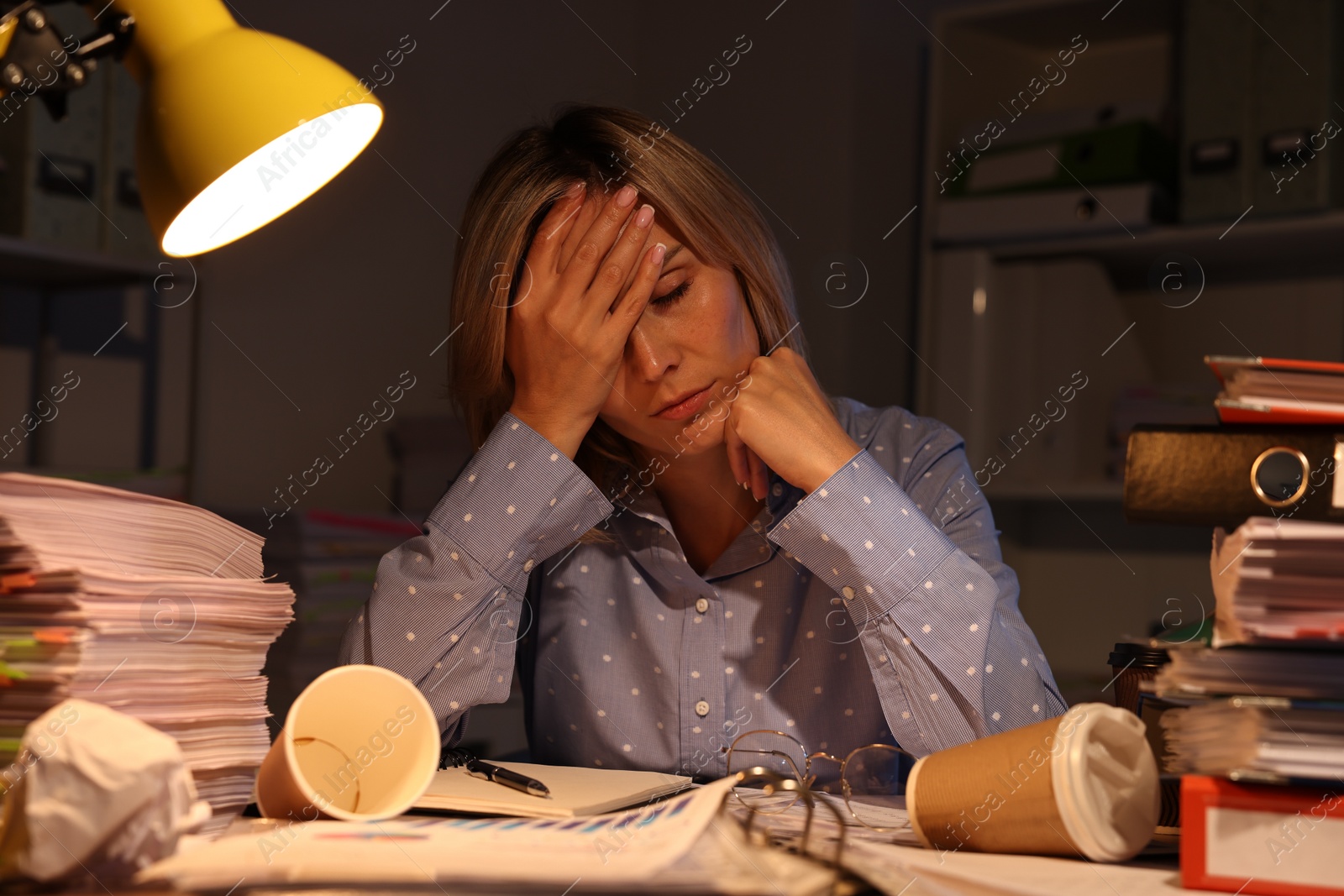 Photo of Overwhelmed woman surrounded by documents and paper coffee cups at table in office at night