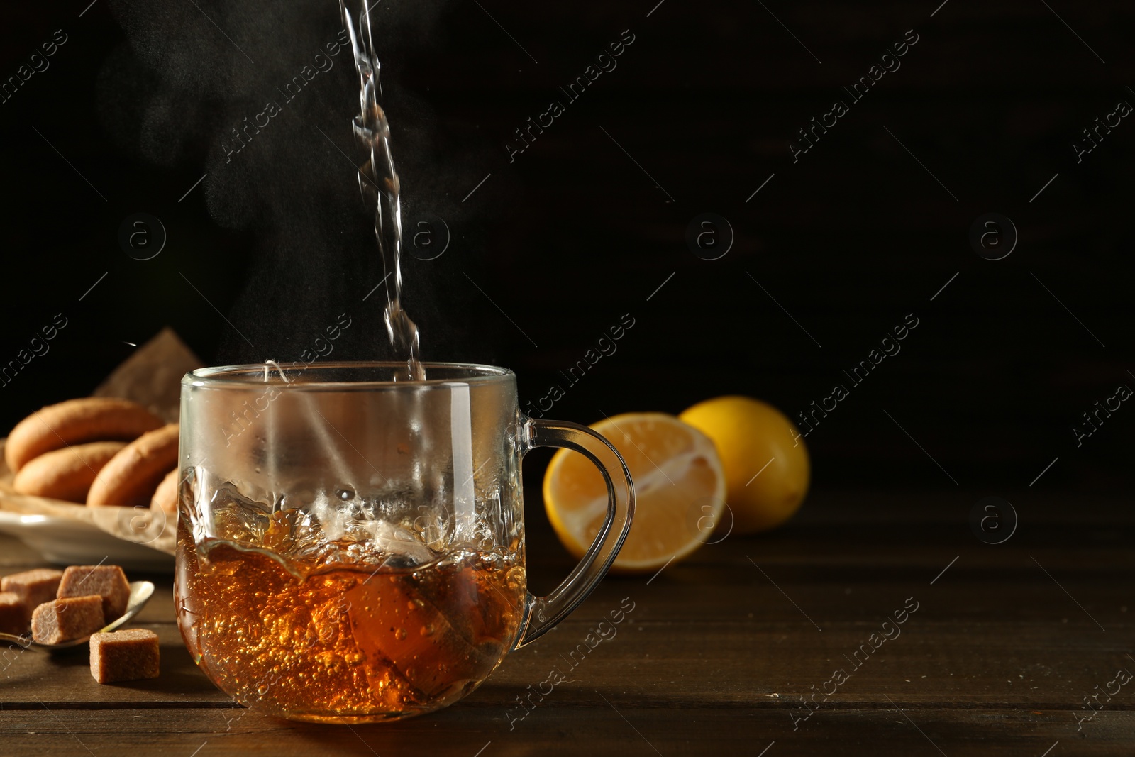 Photo of Pouring hot water into glass cup with tea bag at dark wooden table. Space for text