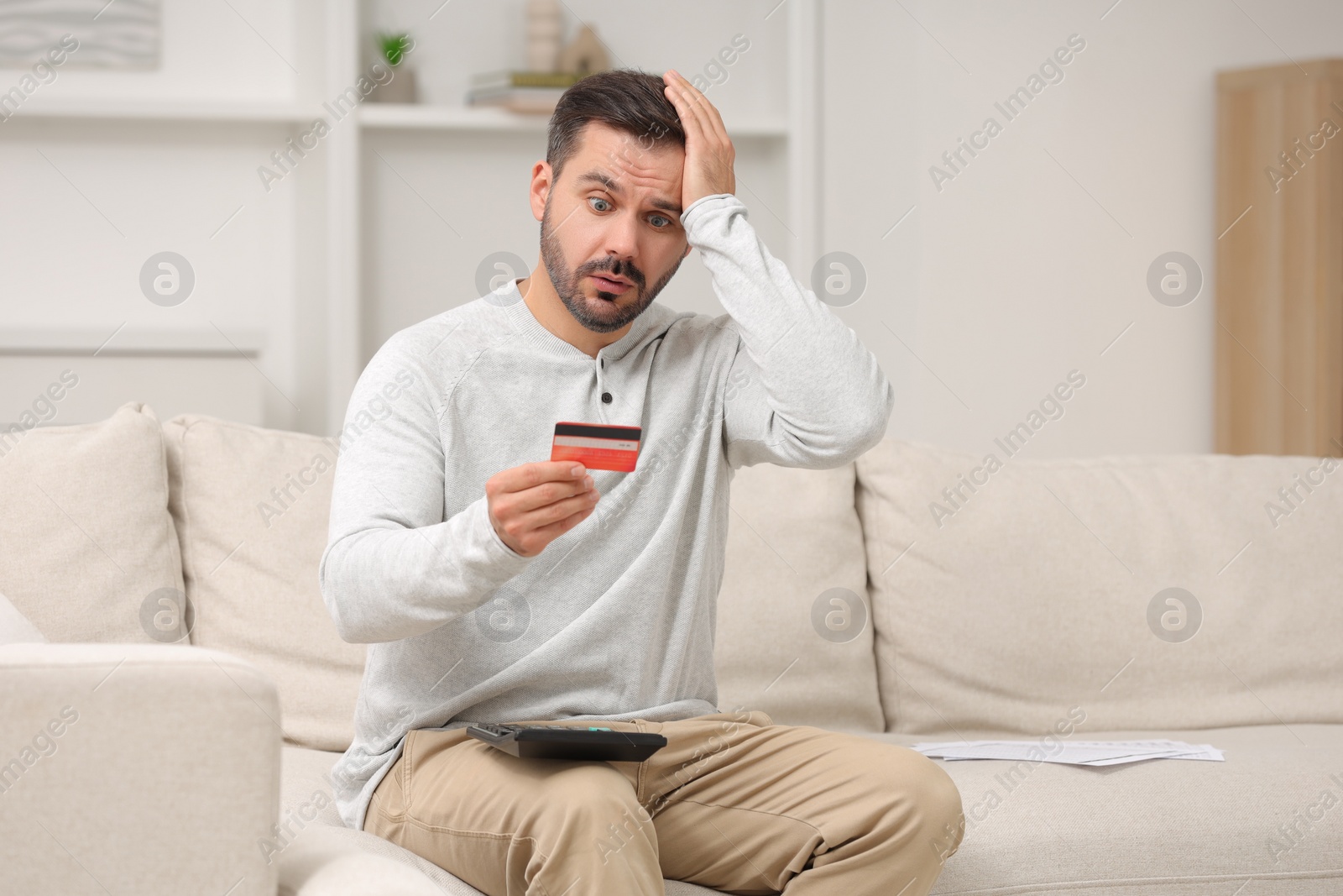 Photo of Shocked man with calculator and credit card planning budget at home. Debt problem