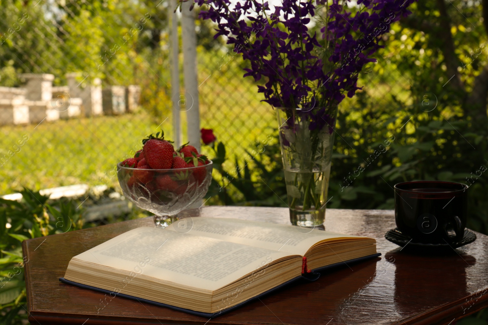 Photo of Open book, cup of tea, strawberries and beautiful wildflowers on table in garden