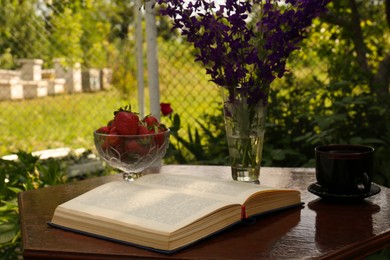 Photo of Open book, cup of tea, strawberries and beautiful wildflowers on table in garden