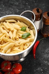 Cooked pasta in metal colander, products and spices on dark textured table, closeup