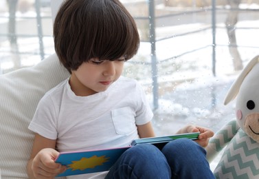 Photo of Cute little boy with toy reading book near window at home