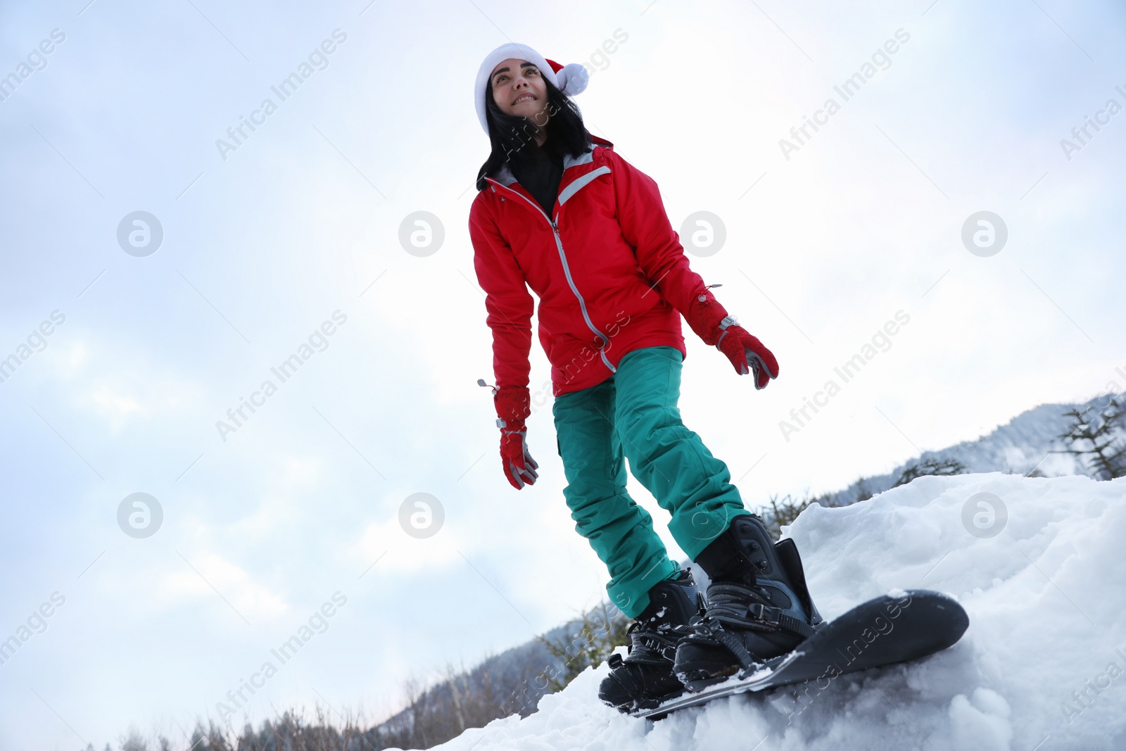 Photo of Young snowboarder wearing Santa hat on snowy hill, low angle view. Winter vacation