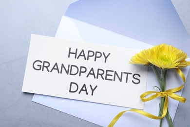 Photo of Card with phrase Happy Grandparents Day, envelope and beautiful flowers on grey background, flat lay