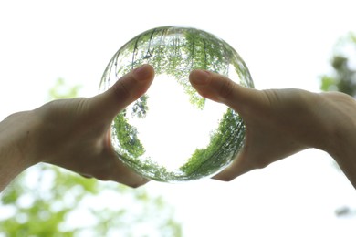 Overturned reflection of beautiful green trees outdoors, low angle view. Man holding crystal ball in park, closeup