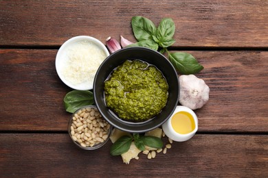 Photo of Tasty pesto sauce in bowl, basil, pine nuts, cheese, garlic and oil on wooden table, top view