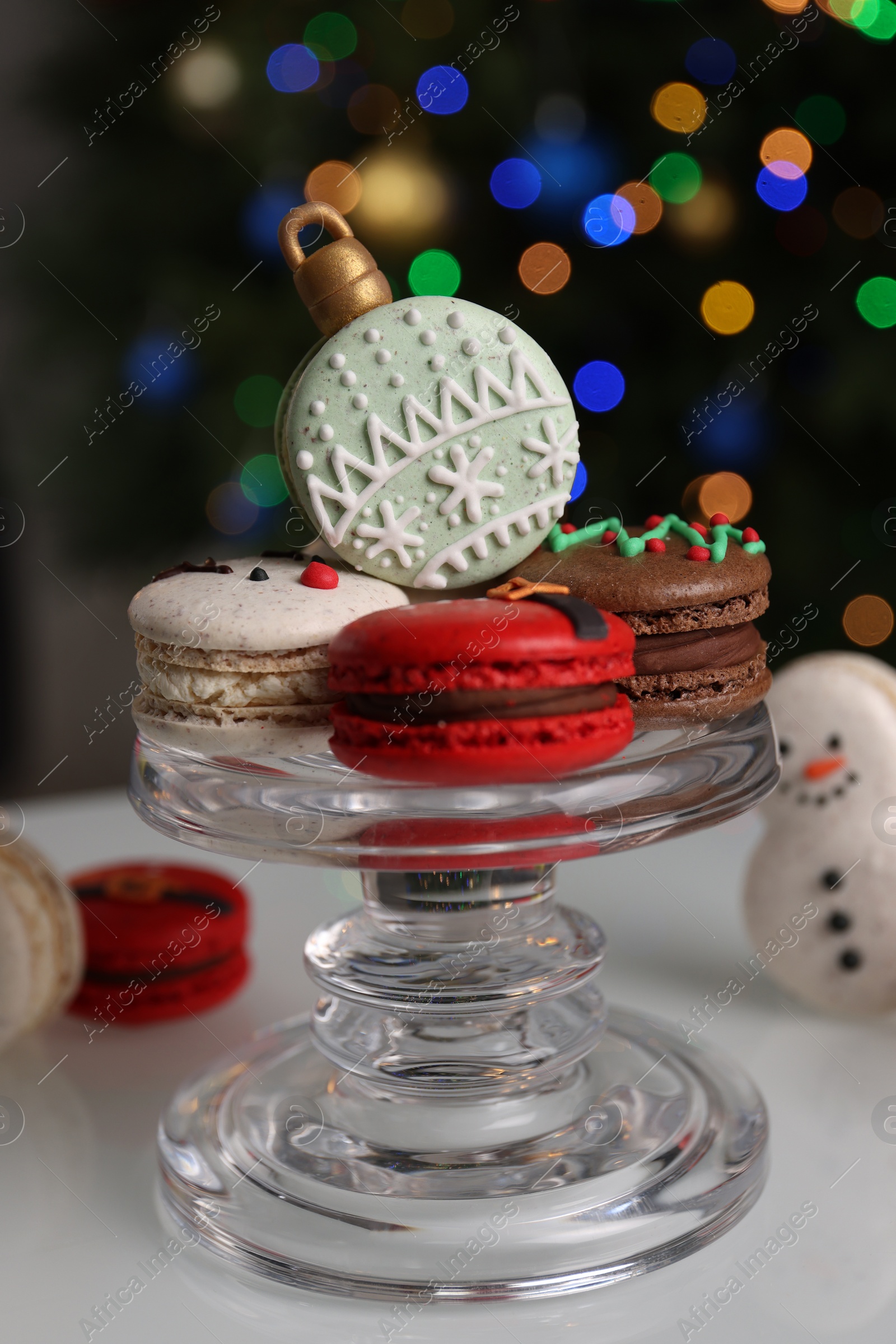 Photo of Beautifully decorated Christmas macarons on white table against blurred festive lights