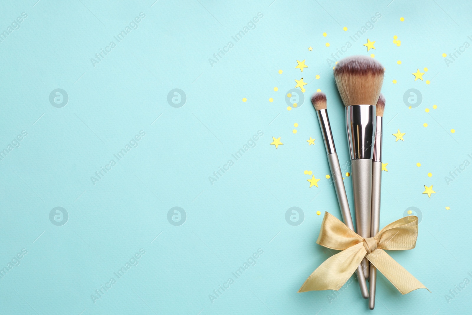 Photo of Different makeup brushes with bow and shiny confetti on turquoise background, flat lay. Space for text