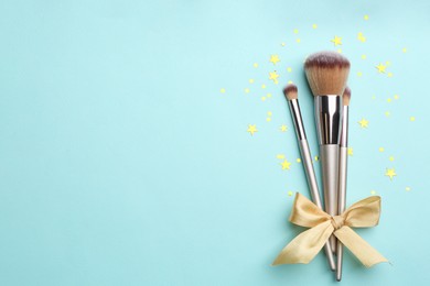 Photo of Different makeup brushes with bow and shiny confetti on turquoise background, flat lay. Space for text