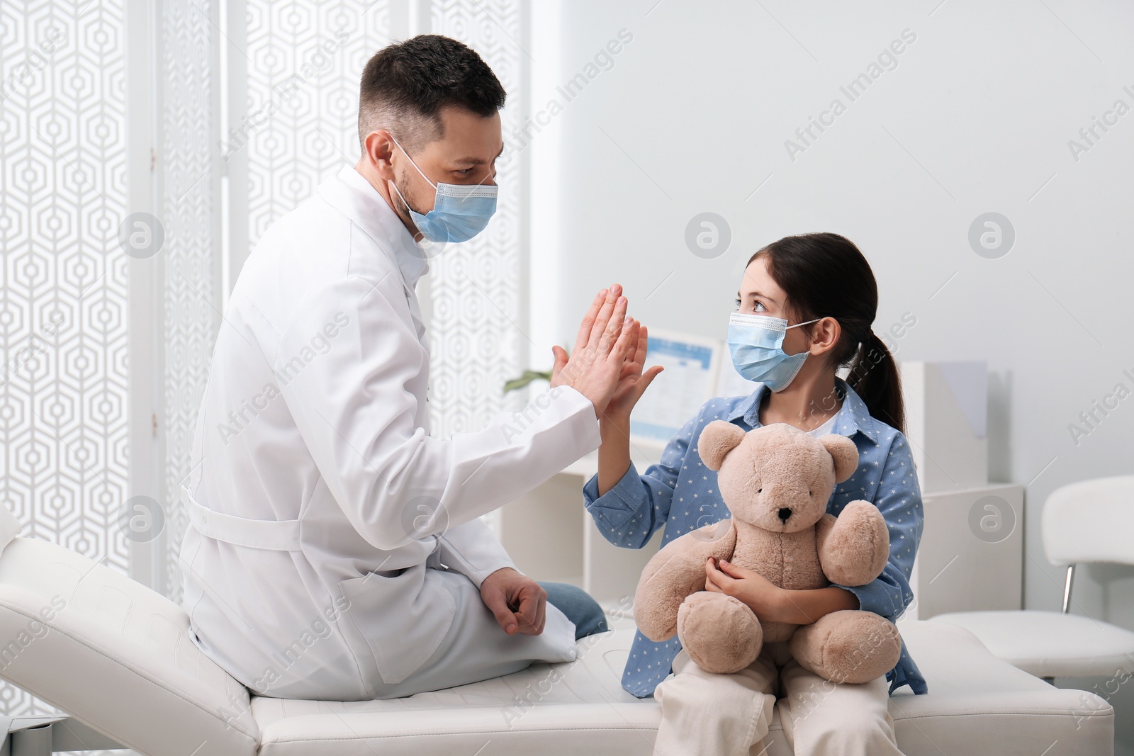 Photo of Pediatrician giving high five to little girl in hospital. Doctor and patient wearing protective masks