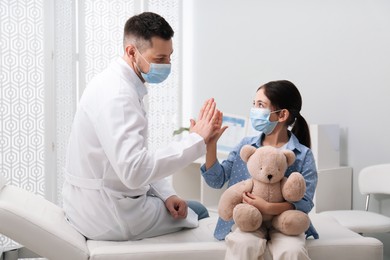 Photo of Pediatrician giving high five to little girl in hospital. Doctor and patient wearing protective masks