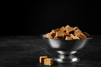 Photo of Metal bowl with brown sugar cubes on black table. Space for text