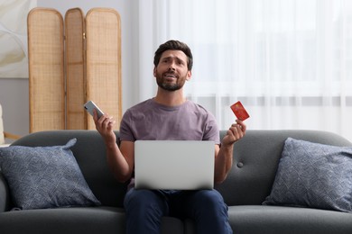 Emotional man with credit card, smartphone and laptop on sofa at home. Be careful - fraud