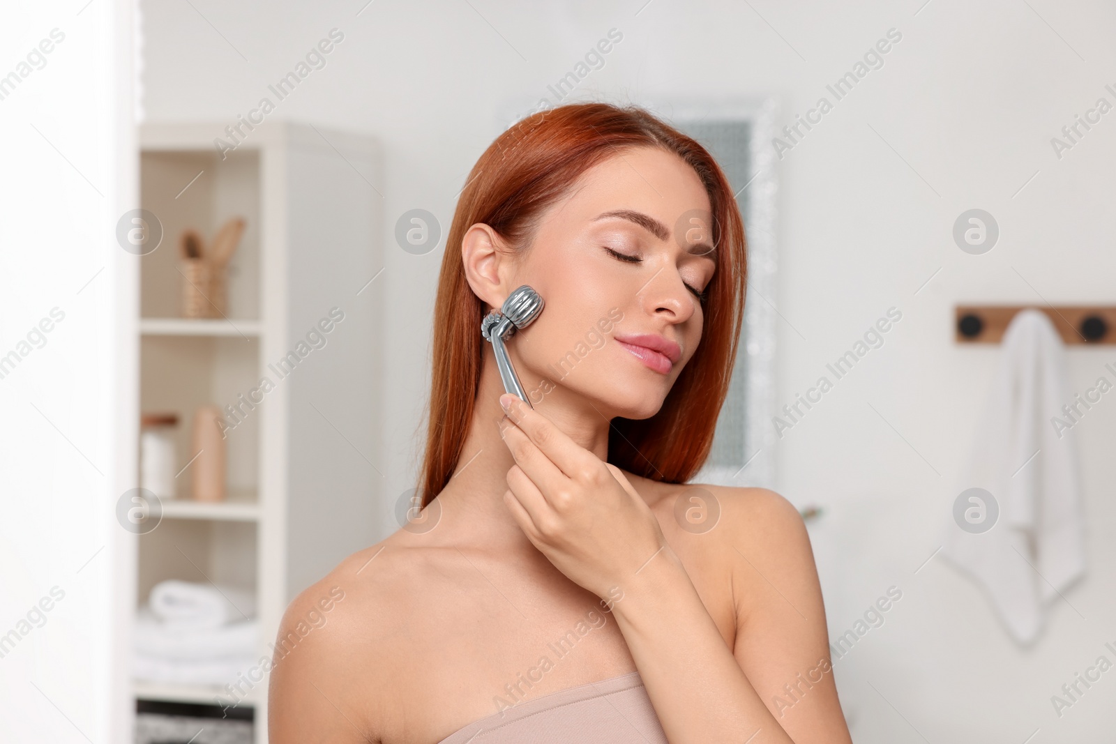 Photo of Young woman massaging her face with metal roller in bathroom