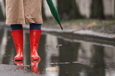 Woman in rubber boots with umbrella walking outdoors on rainy day, closeup. Space for text