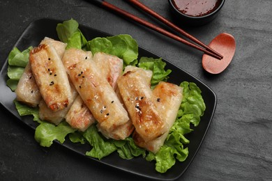 Photo of Tasty fried spring rolls, lettuce and sauce on black table, top view