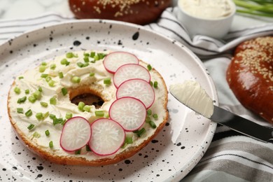 Photo of Delicious bagel with cream cheese, green onion and radish on plate, closeup
