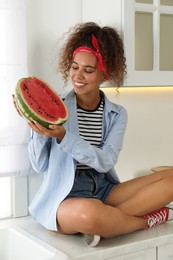 Photo of Beautiful young African American woman with half of watermelon sitting on countertop in kitchen