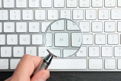 Photo of Woman holding magnifier glass over keyboard on black slate table, top view. Find keywords concept