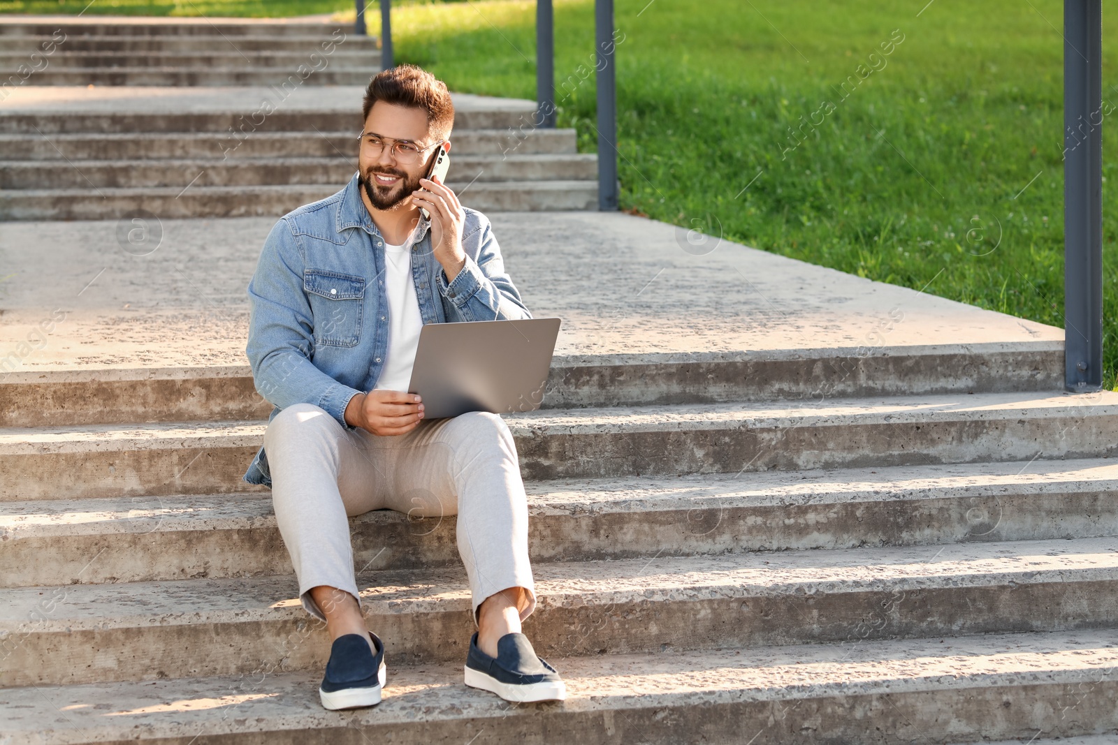 Photo of Handsome young man with laptop talking on smartphone on concrete stairs outdoors. Space for text