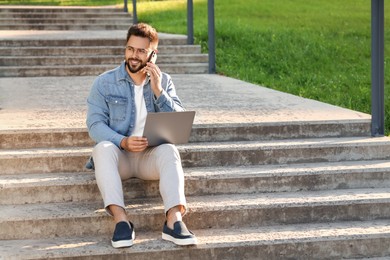 Handsome young man with laptop talking on smartphone on concrete stairs outdoors. Space for text
