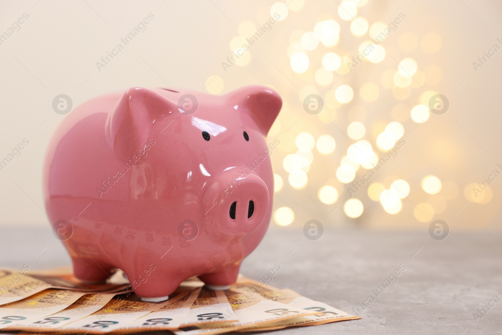 Photo of Piggy bank with euro banknotes on grey table against blurred lights, space for text